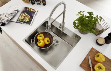 Moderne Edelstahlspüle mit gelben Paprikas im Sieb, frischer Petersilie und Kochbuch auf weißer Kücheninsel
